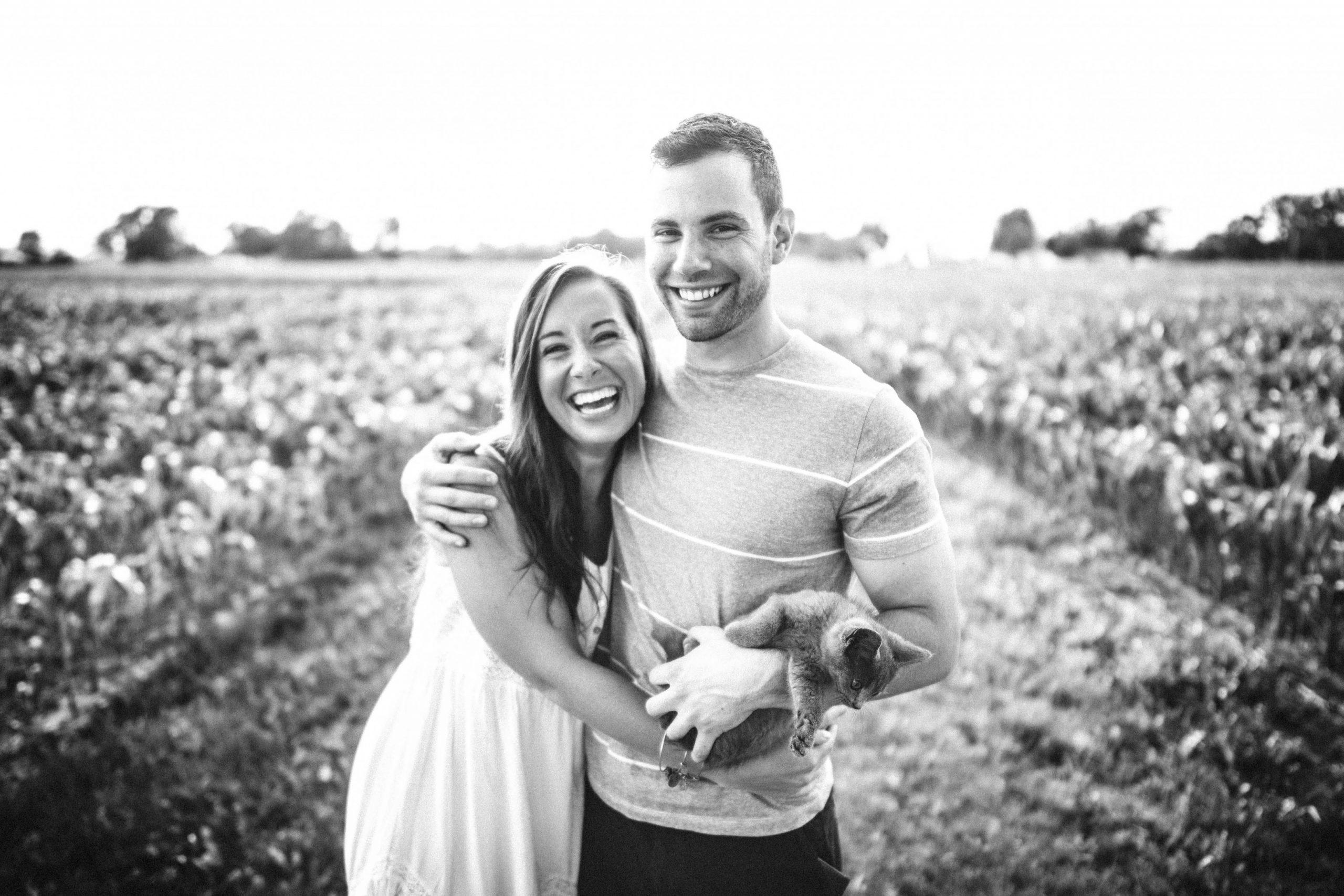 black and white image of young smiling couple with cat in field scaled